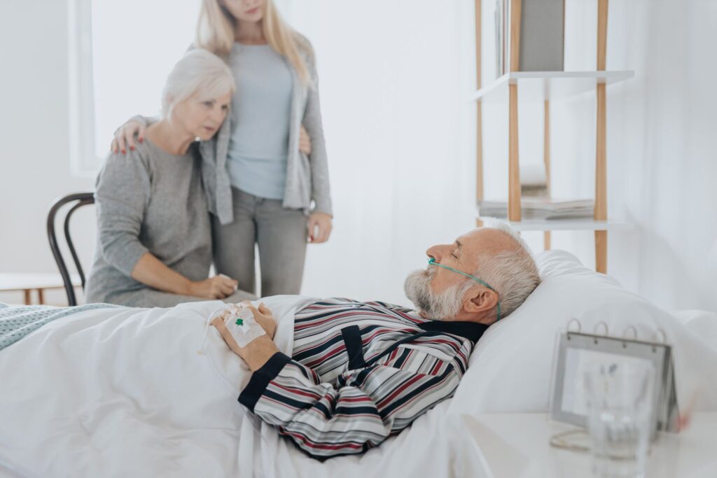 Zwei Damen, hypothetisch Mutter und Tochter, stehen neben einem älteren, kranken Herrn, der ans Bett gefesselt ist.